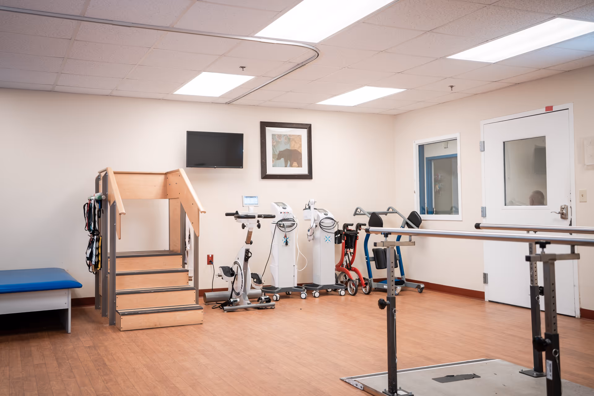 Bright indoor rehab room with parallel bars, exercise machines, and a wooden staircase.