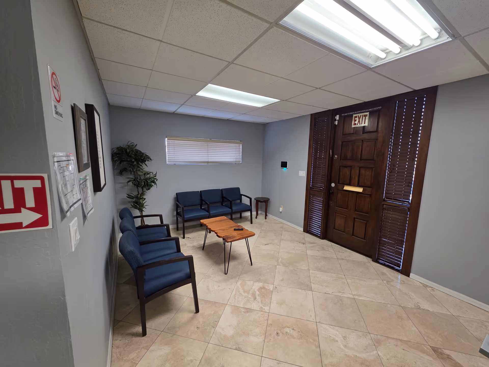 A small lobby with blue chairs, a wooden coffee table, plant, and exit door.