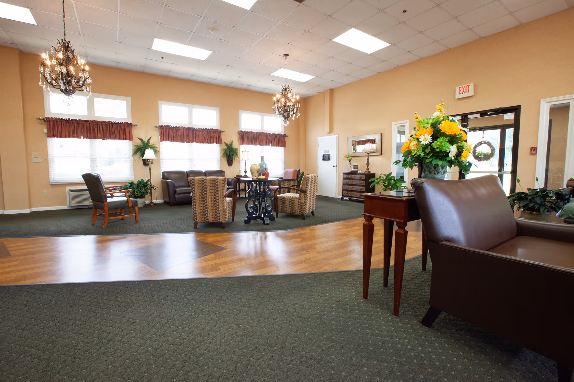 A spacious and well-lit common area in a senior living facility with comfortable seating including leather chairs and patterned armchairs arranged around small tables. The room features large windows with red valances, green plants, chandeliers, and a mix of carpet and wood flooring. A table with a large floral arrangement is visible in the foreground near the entrance door.