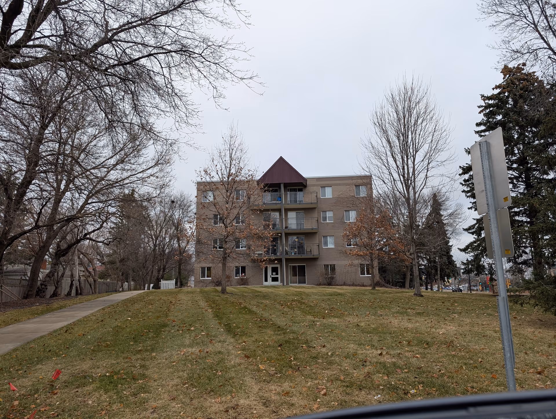 Four-story brick apartment building with balconies seen from a grassy front lawn lined with leafless trees and a sidewalk.