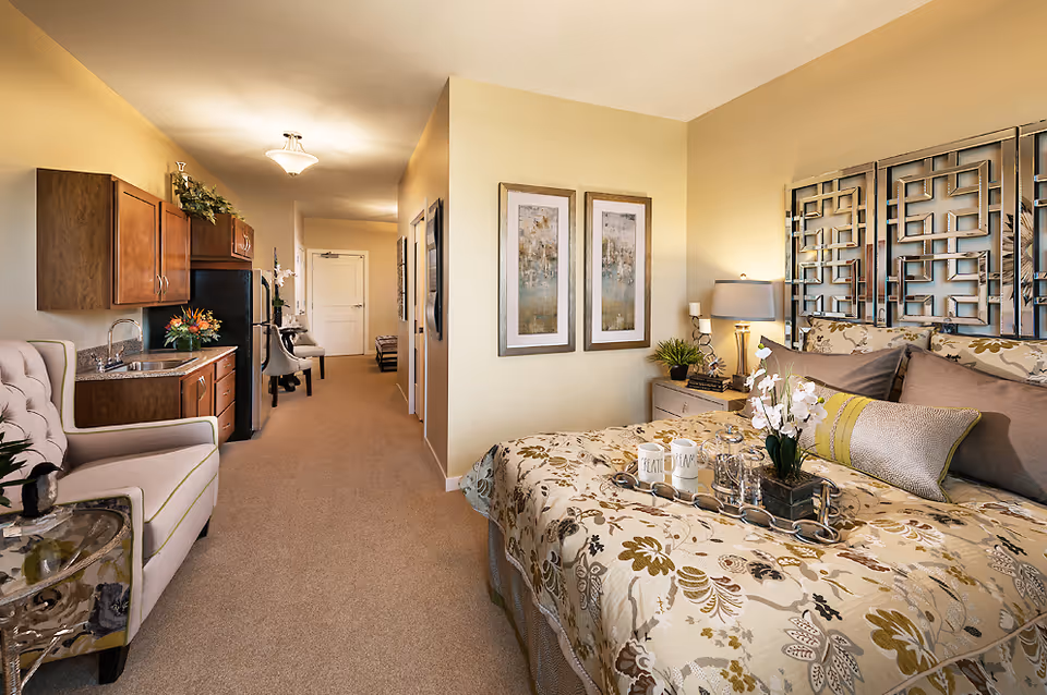 Well-lit senior living suite with a patterned bed, nightstands, and a compact kitchen area.