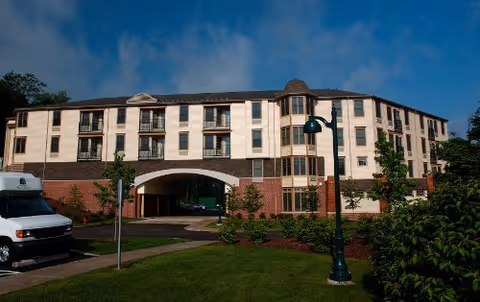 Three-story beige senior living building with brick lower level, archway, driveway, and lawn.
