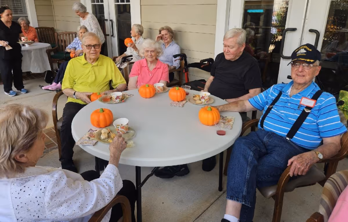 Elderly residents sit around a round table with pumpkins on an outdoor patio.