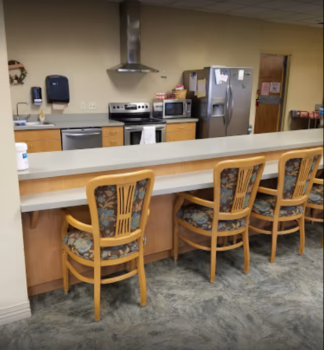 Communal kitchen with a long counter, wood-framed floral chairs, and appliances along the back wall.