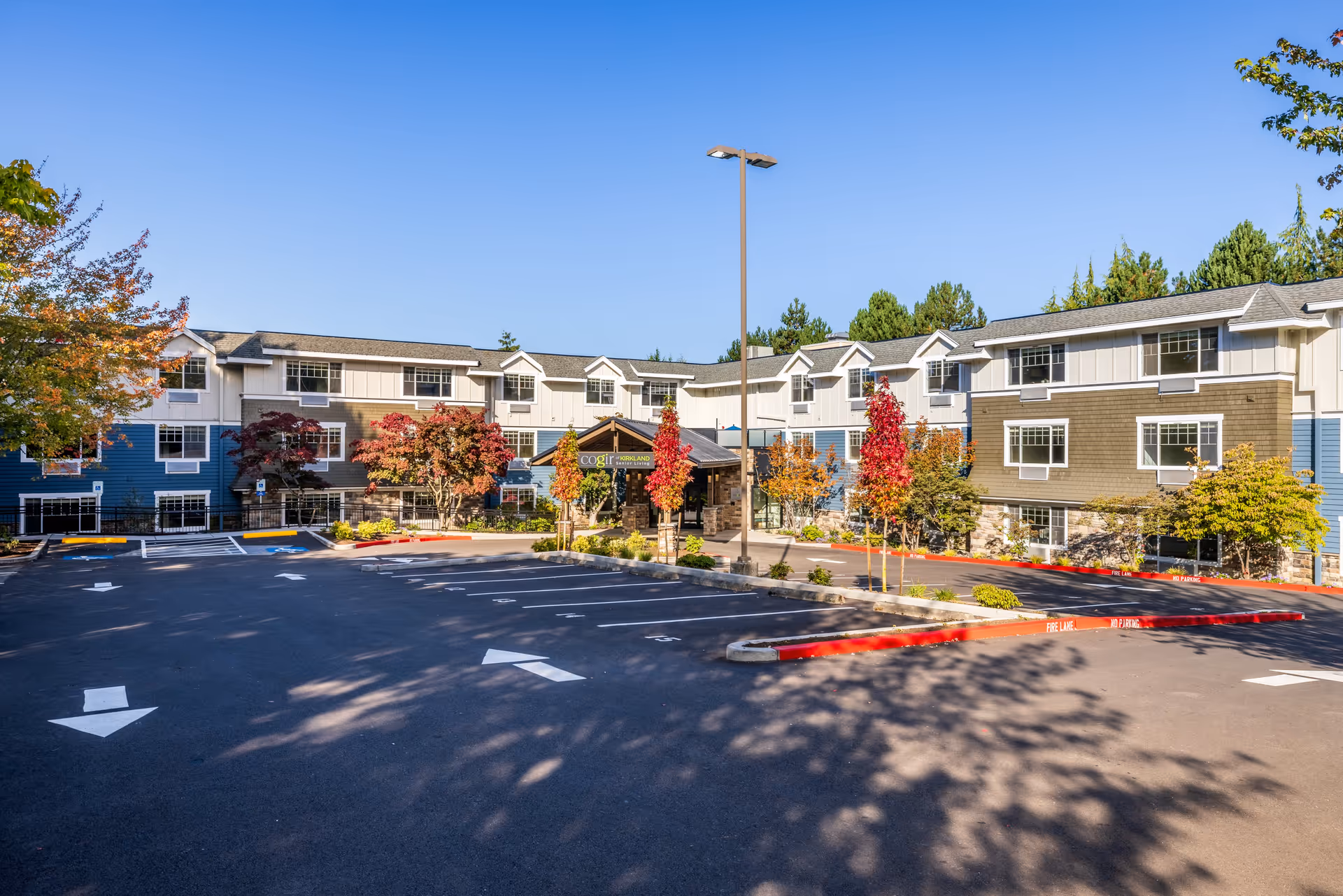Exterior view of COGIR Senior Living facility showing a three-story building with a mix of blue, white, and brown siding, surrounded by trees with autumn foliage. The foreground features an empty parking lot with directional arrows and a tall street lamp.
