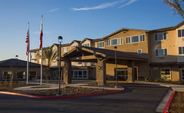 Exterior view of Renaissance Village Murrieta, showing a multi-story building with a covered entrance, flagpoles with flags, and a driveway under a clear blue sky.