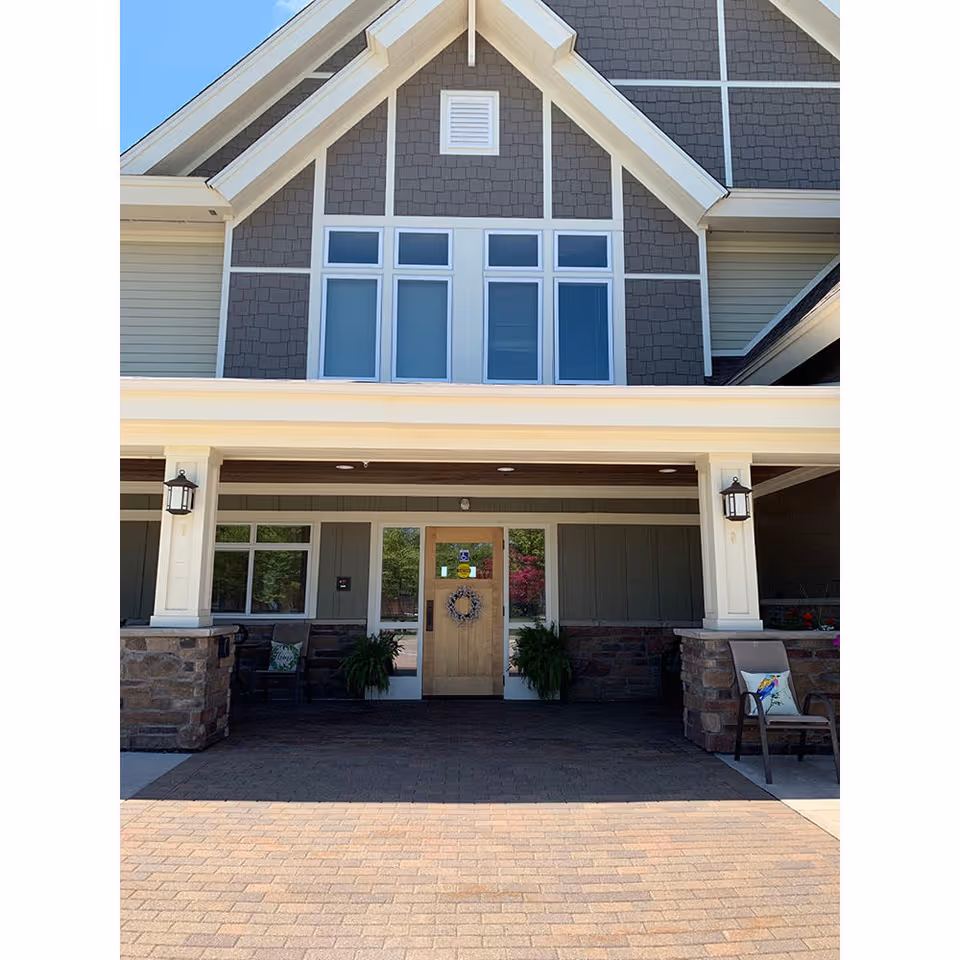 Front entrance of a building with a peaked roof and multiple windows above the door. The entrance has a covered porch supported by columns with stone bases. There are two chairs with decorative pillows on either side of the door, and two potted plants flanking the door which has a wreath hanging on it.