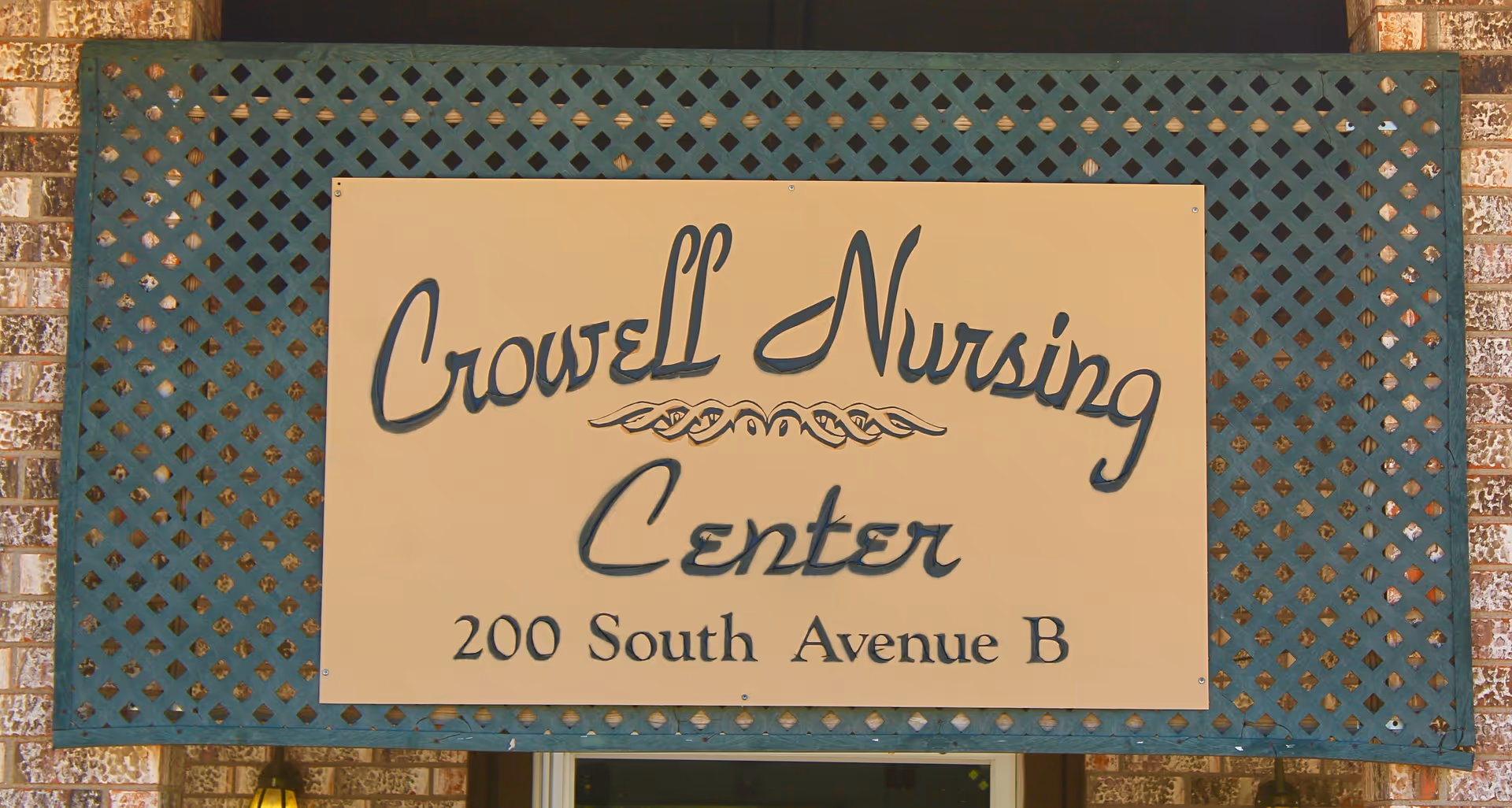 A sign mounted on a green lattice background with brick walls on either side. The sign reads 'Crowell Nursing Center 200 South Avenue B' in decorative black lettering on a beige background.