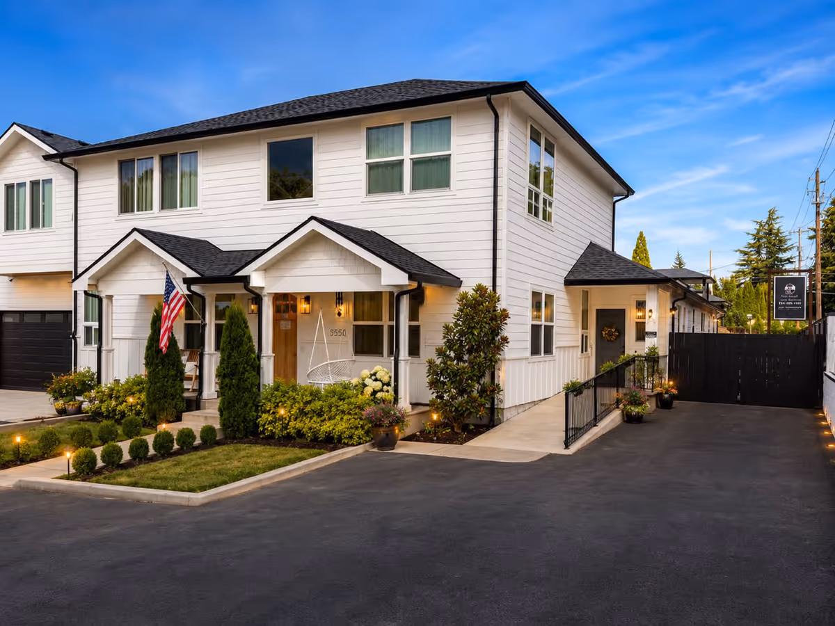 White two story senior living facility with front porch, American flag, and well kept landscaping.