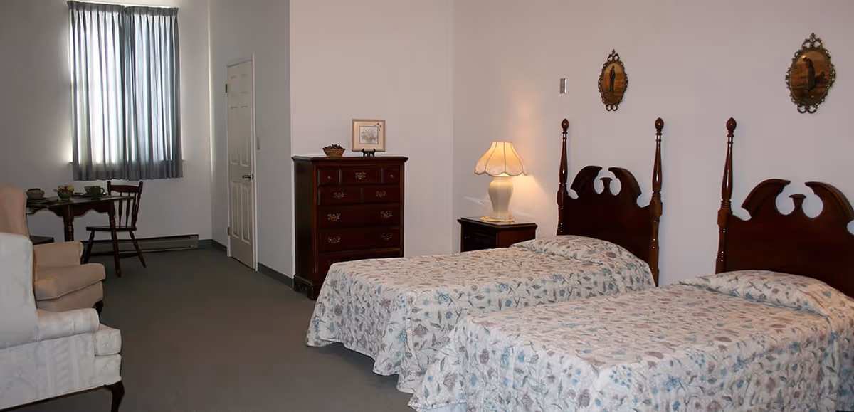 A bedroom with two twin beds featuring wooden headboards and floral bedspreads. There is a nightstand with a lamp between the beds, a wooden dresser against the wall, a small table with two chairs near a window with closed curtains, and an upholstered armchair in the foreground.