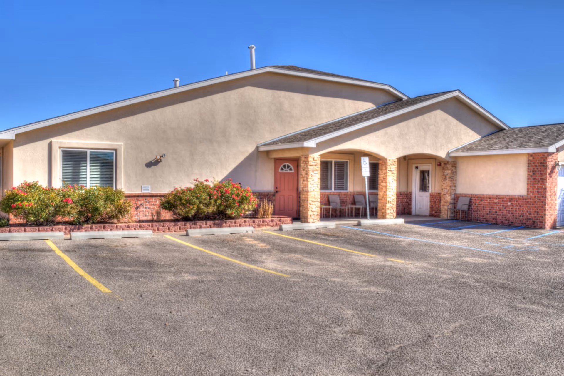 Beige assisted living building with stone pillars, entrance, and empty parking lot under blue sky.
