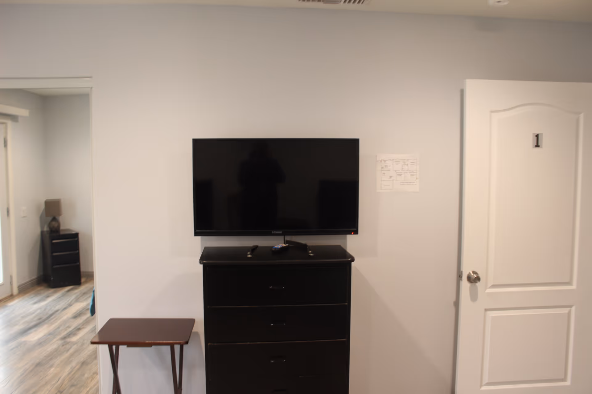 TV on black dresser with small table in neutral interior, doorway labeled 1 nearby.