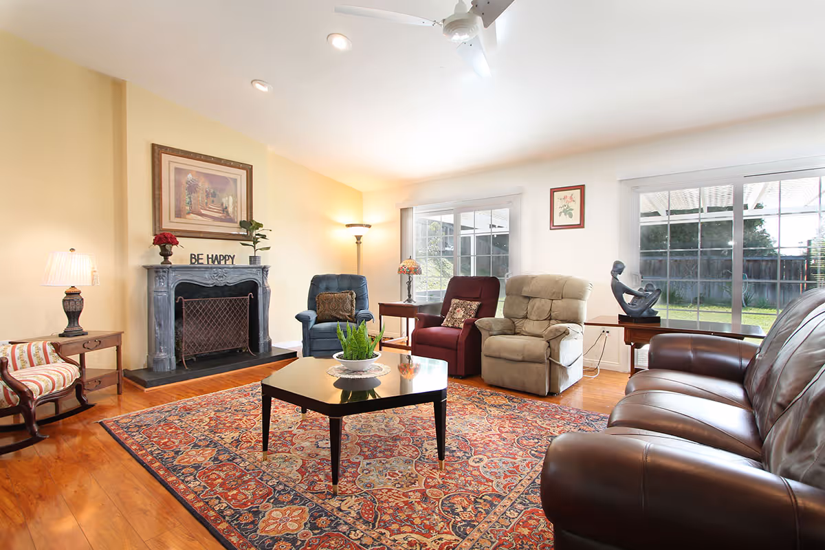 A bright and cozy living room with a wooden floor and a large patterned rug. The room features a dark leather sofa, three armchairs in blue, red, and beige, a black coffee table with a plant centerpiece, a fireplace with a 'BE HAPPY' sign on the mantel, a floor lamp, and large windows showing a green backyard.