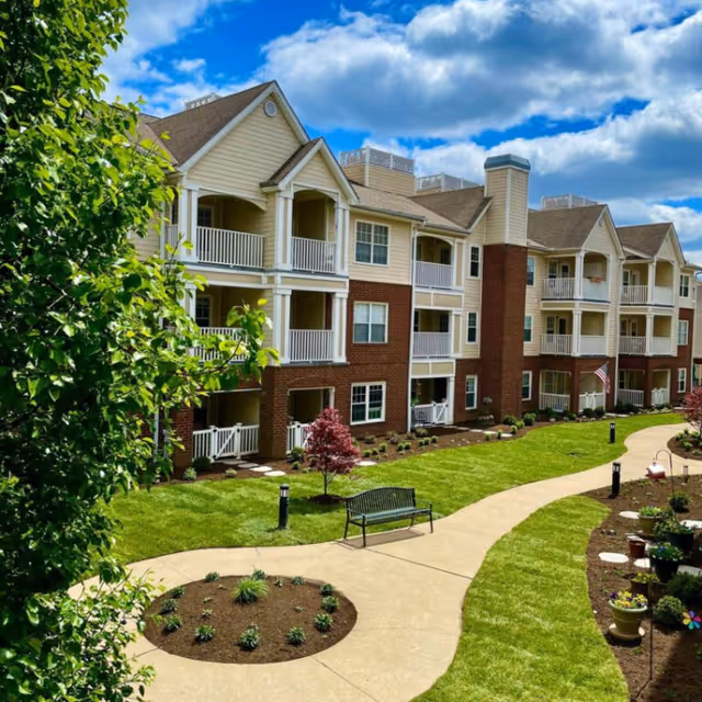 Exterior view of a multi-story senior living facility with balconies, a well-maintained lawn, landscaped garden beds, a curved concrete walkway, a bench, and a partly cloudy blue sky.