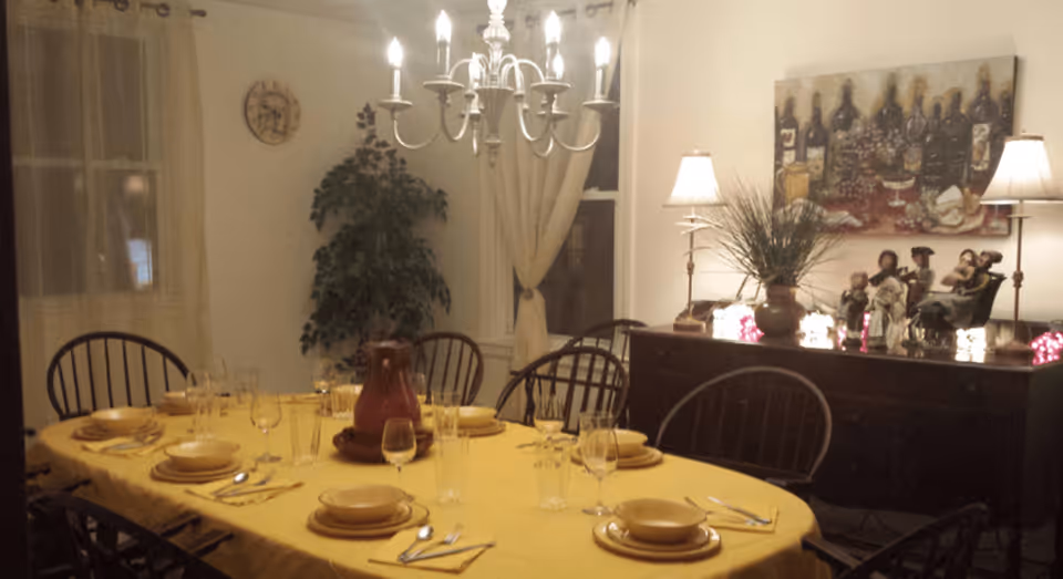 A warmly lit dining room with a long table covered by a yellow tablecloth, set with plates, bowls, glasses, and silverware for eight people. A red pitcher is placed in the center of the table. The room features a chandelier overhead, a large plant near the window with curtains, and a sideboard decorated with lamps, figurines, a potted plant, and a painting of wine bottles and grapes on the wall.