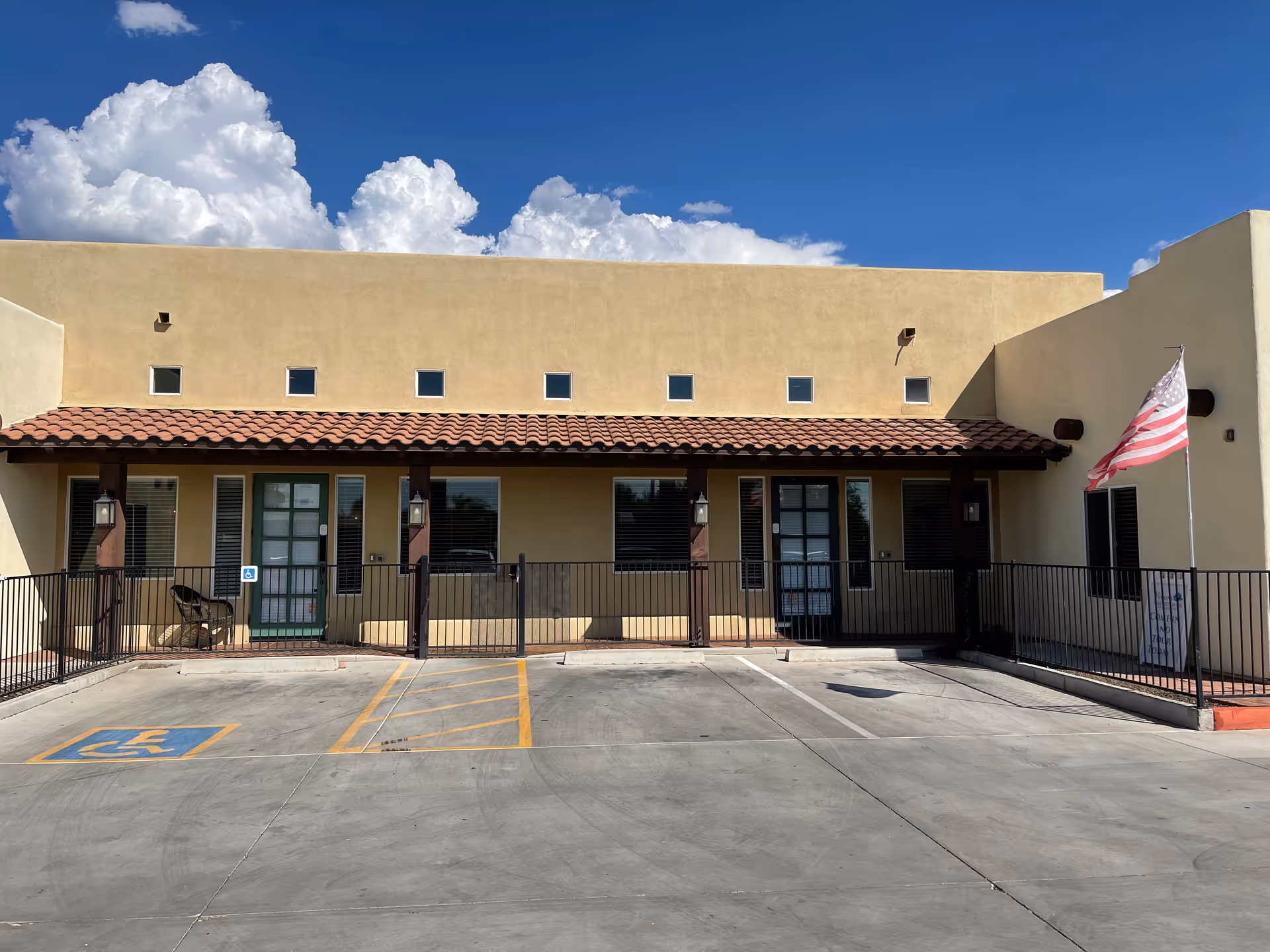 Single-story tan building with a red-tile roof, railing, handicap space, and American flag in front.