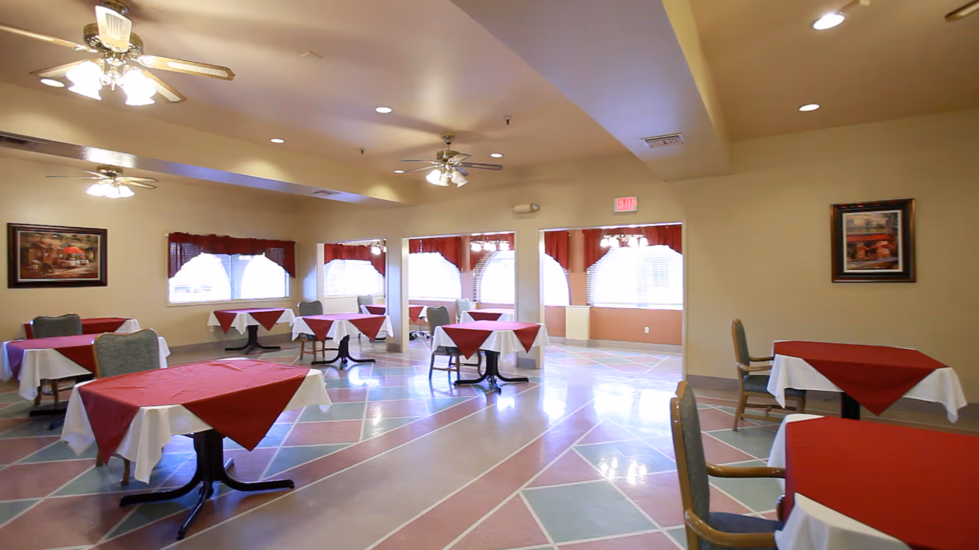 A dining room with multiple tables covered with white tablecloths and red overlays. Each table is paired with chairs, and the room features ceiling fans with lights. The floor has a geometric pattern with pastel colors, and there are windows with red valances letting in natural light. Two framed pictures hang on the beige walls.