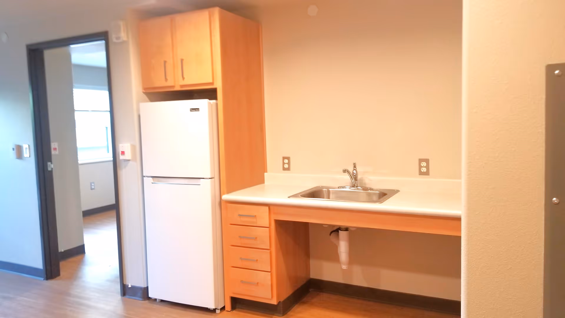 A small kitchen area with a white refrigerator, wooden cabinets, a countertop with a stainless steel sink, and an open space underneath the sink. The kitchen is adjacent to a hallway leading to another room with a window.