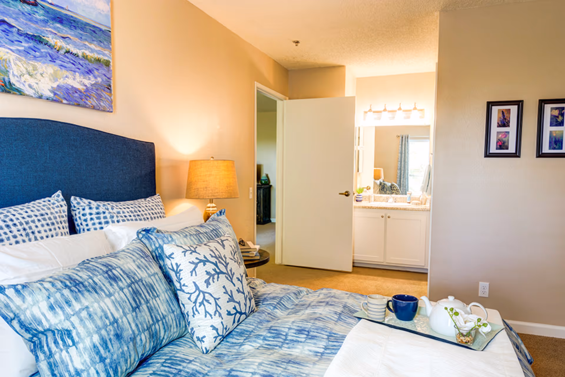 A cozy bedroom with a blue upholstered headboard and blue patterned pillows and bedding. A bedside table with a lamp is next to the bed. The door to an adjoining bathroom is open, showing a vanity with a large mirror and bright lights. Two framed pictures hang on the beige wall.