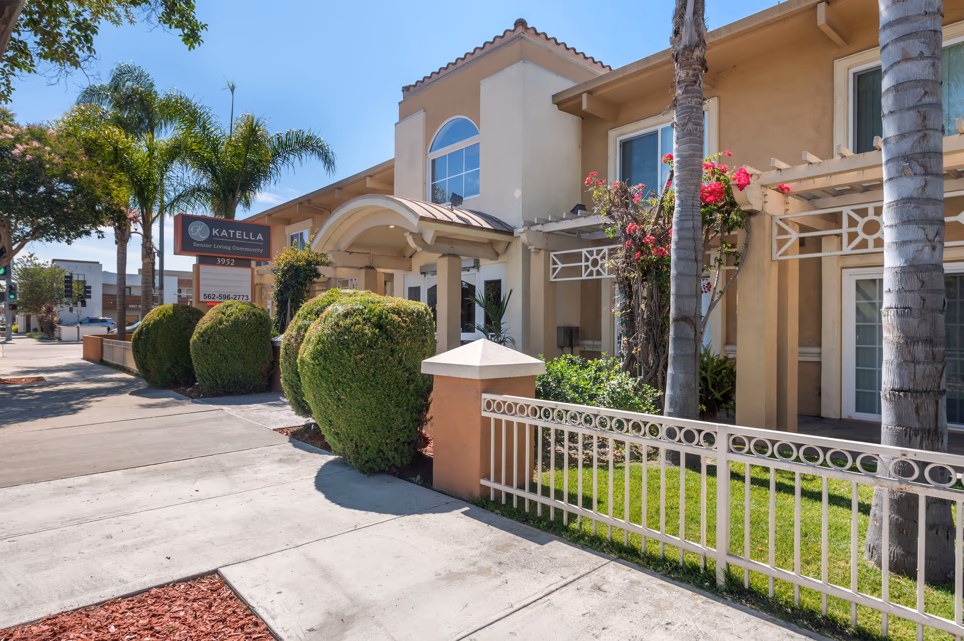 Exterior view of Katella Senior Living Community building with beige walls, arched entrance, trimmed bushes, palm trees, and a sidewalk in front under a clear blue sky.