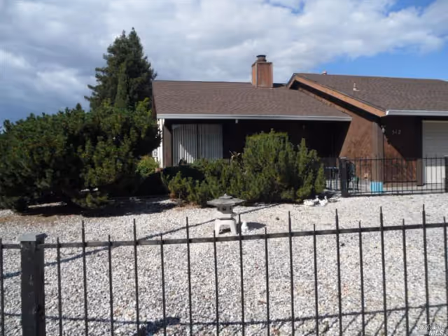 Single-story ranch-style house with a gravel front yard, low metal fence, shrubs, and a covered porch under a partly cloudy sky.