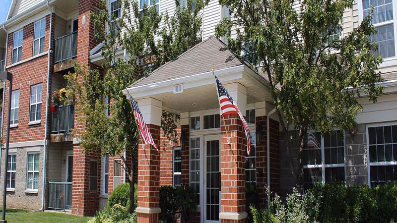 Entrance of a brick senior living building with a covered portico, two American flags, windows, balconies, and landscaping.