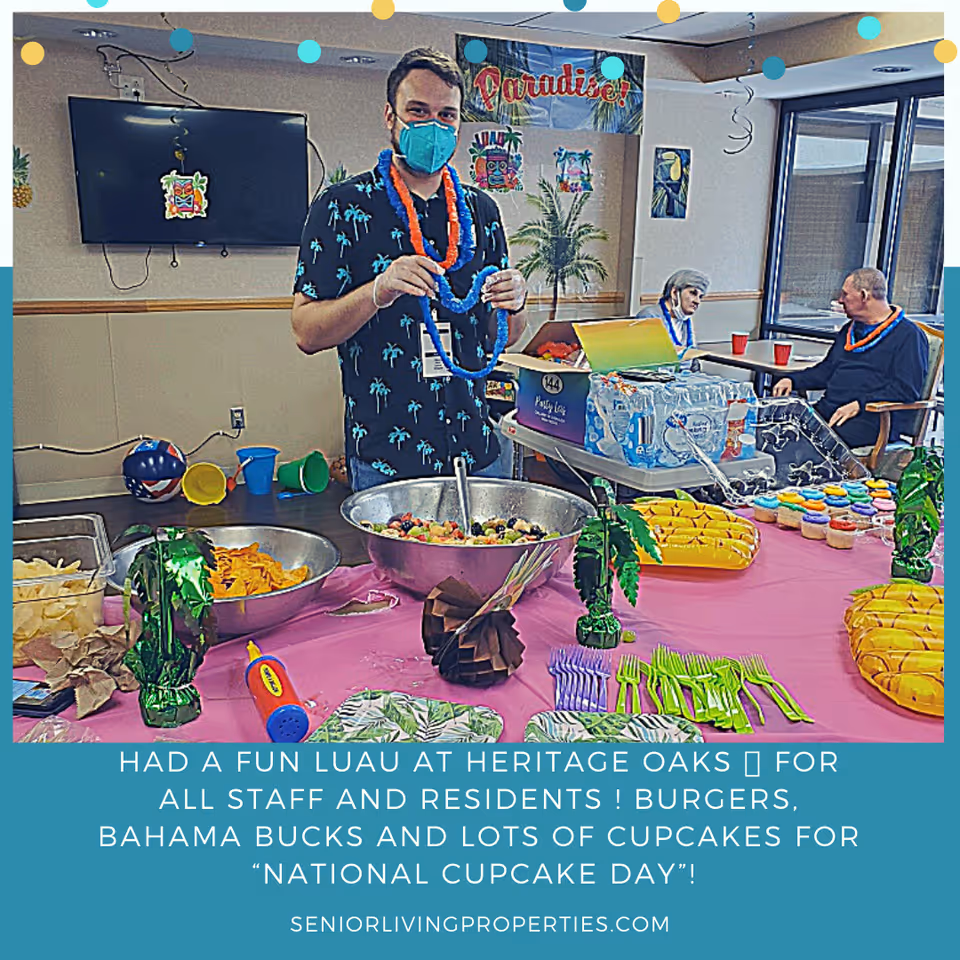 A man wearing a blue face mask and a black shirt with palm tree prints stands behind a table with bowls of snacks and fruit salad. The table is decorated with tropical-themed items including green palm tree decorations, yellow inflatable bananas, and colorful plastic utensils. In the background, two elderly people wearing leis sit at a table with red cups. The room is decorated with a 'Paradise!' banner and tropical-themed wall decorations.