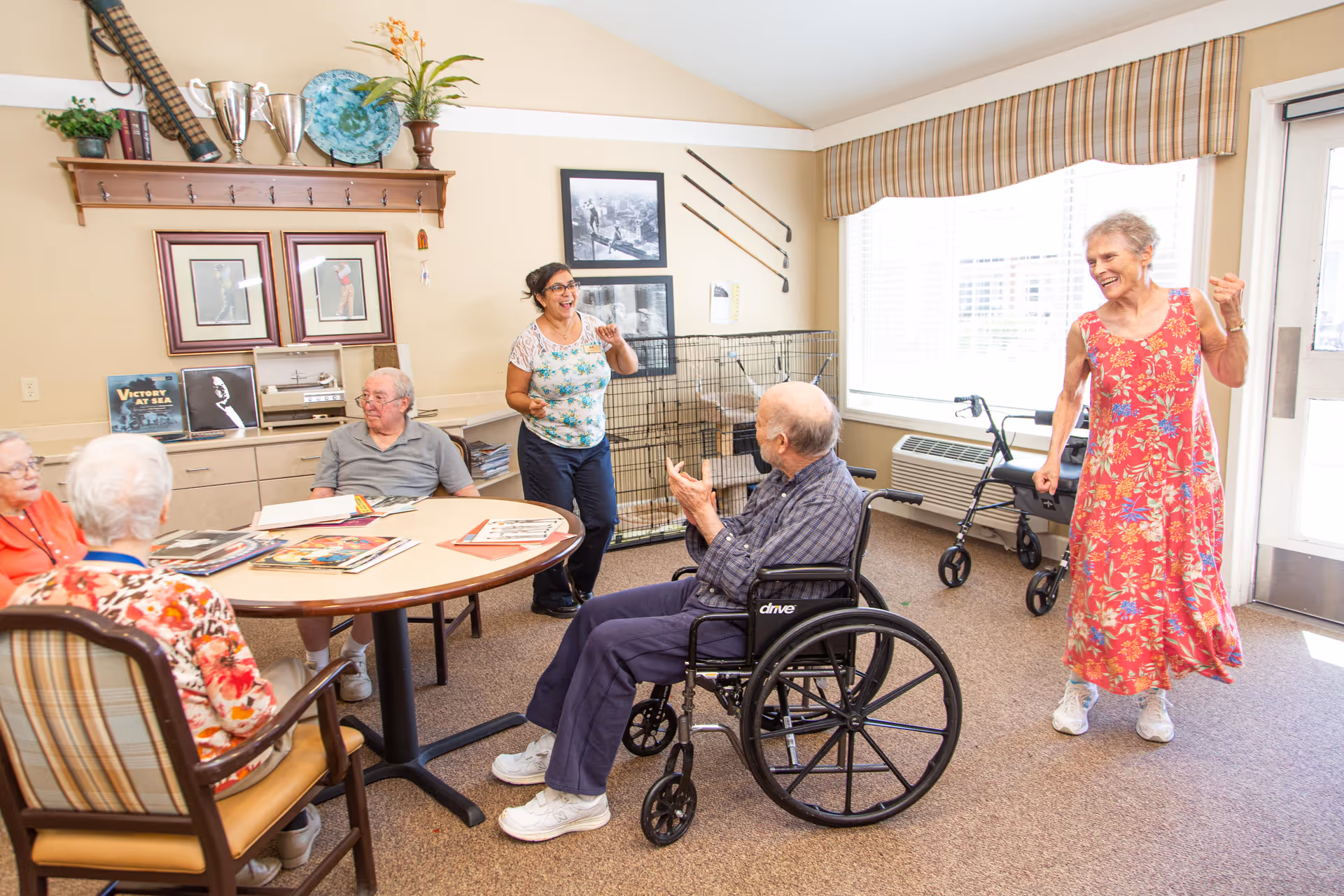 A group of elderly people and a caregiver are gathered in a bright room with beige walls and carpet. Three elderly individuals are seated around a round table with magazines and papers, one man in a wheelchair is clapping, and a woman in a red floral dress is standing and smiling near a window with blinds. The caregiver is standing and engaging with the group. The room has framed pictures on the wall, a shelf with trophies and plants, and a walker near the window.