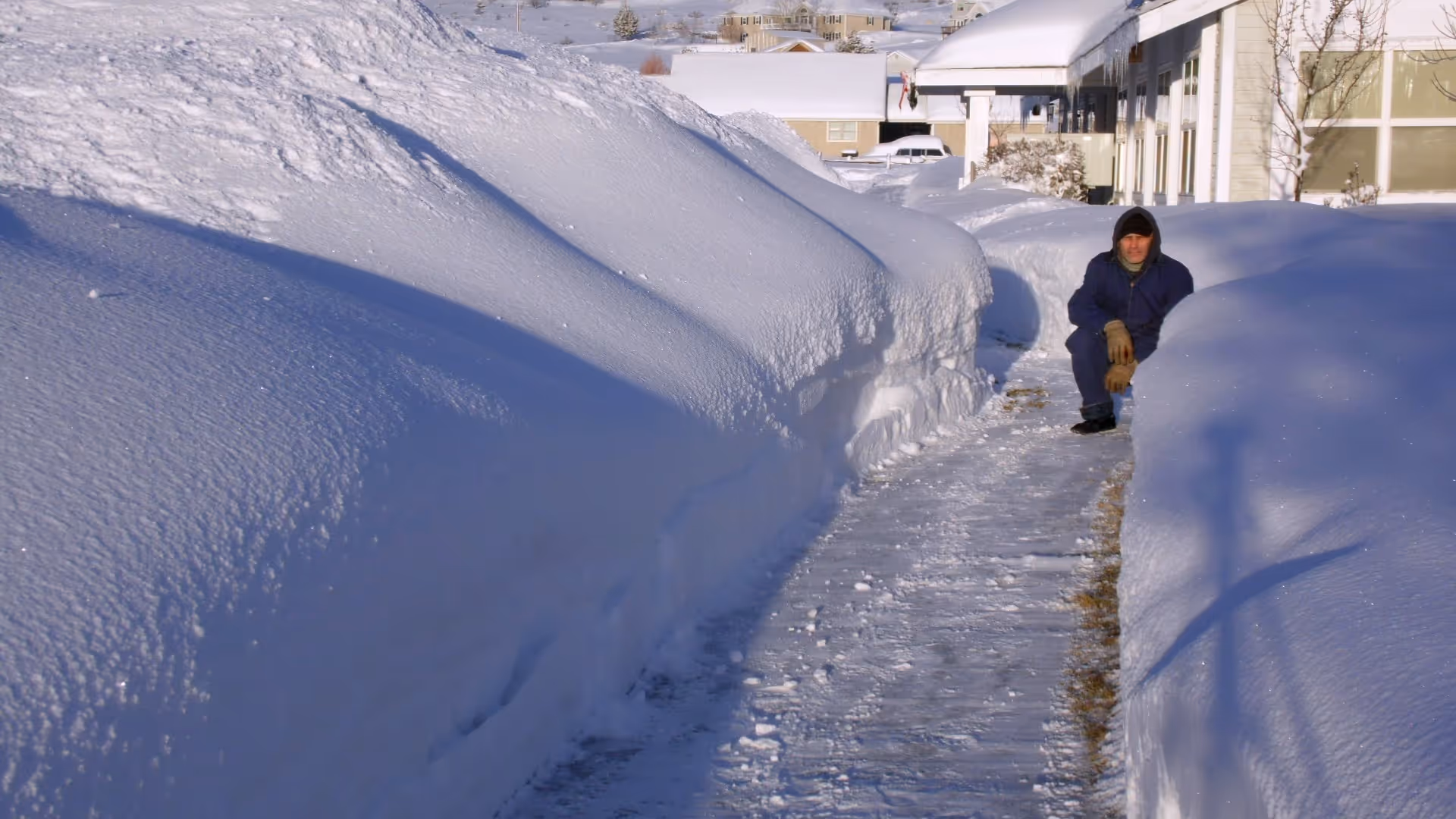 A man in winter gear squats on a cleared outdoor path beside tall snowbanks near buildings.