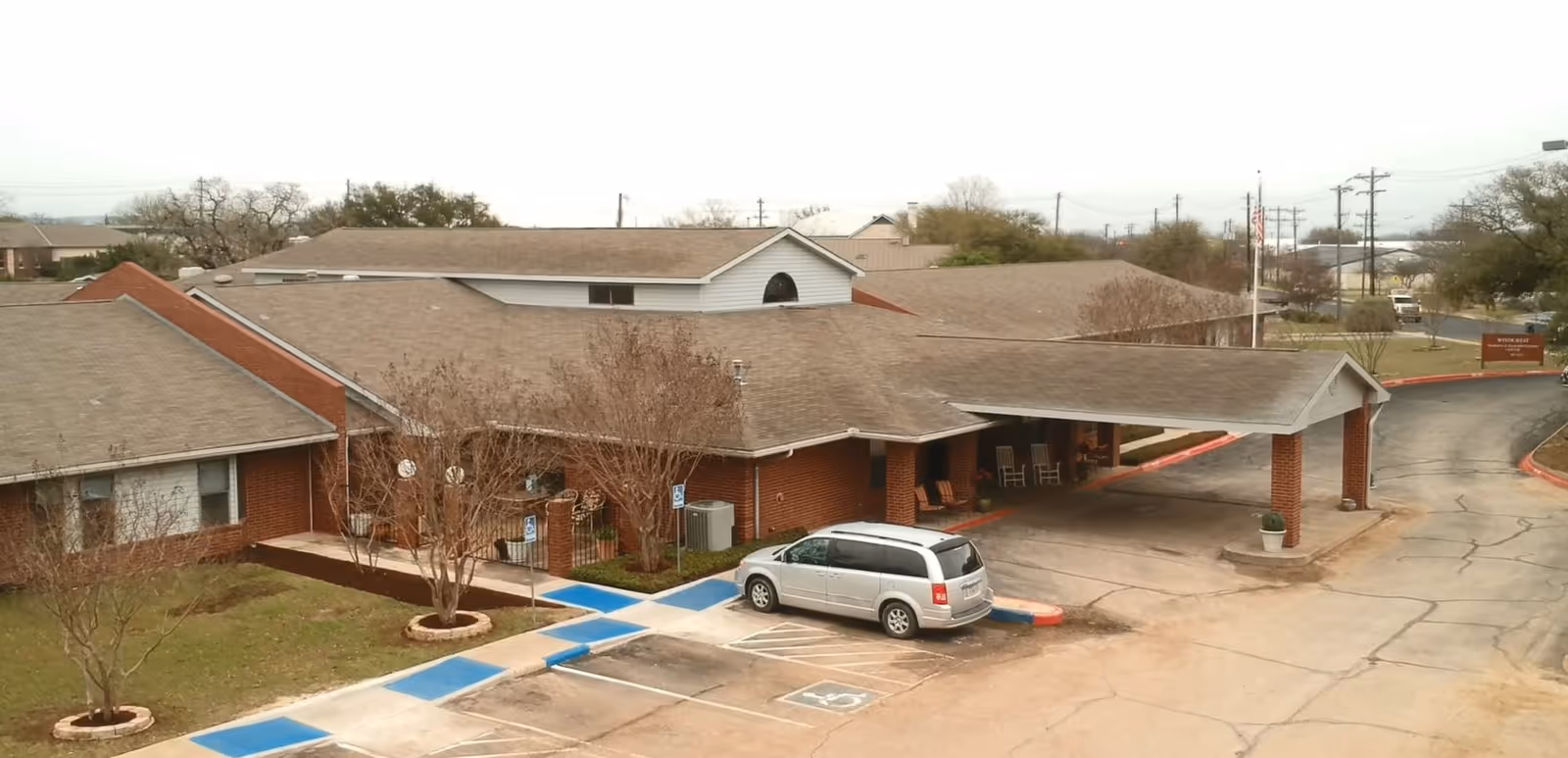Exterior view of a single-story brick building with a covered entrance and a silver minivan parked in a handicapped parking space. The building is surrounded by a paved driveway and some leafless trees, with a sign visible in the background near the road.