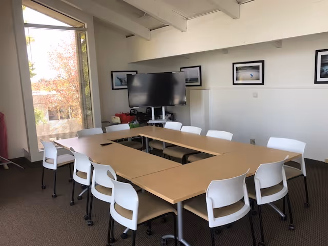 U-shaped tables with white chairs surround a bright meeting room and mounted television.