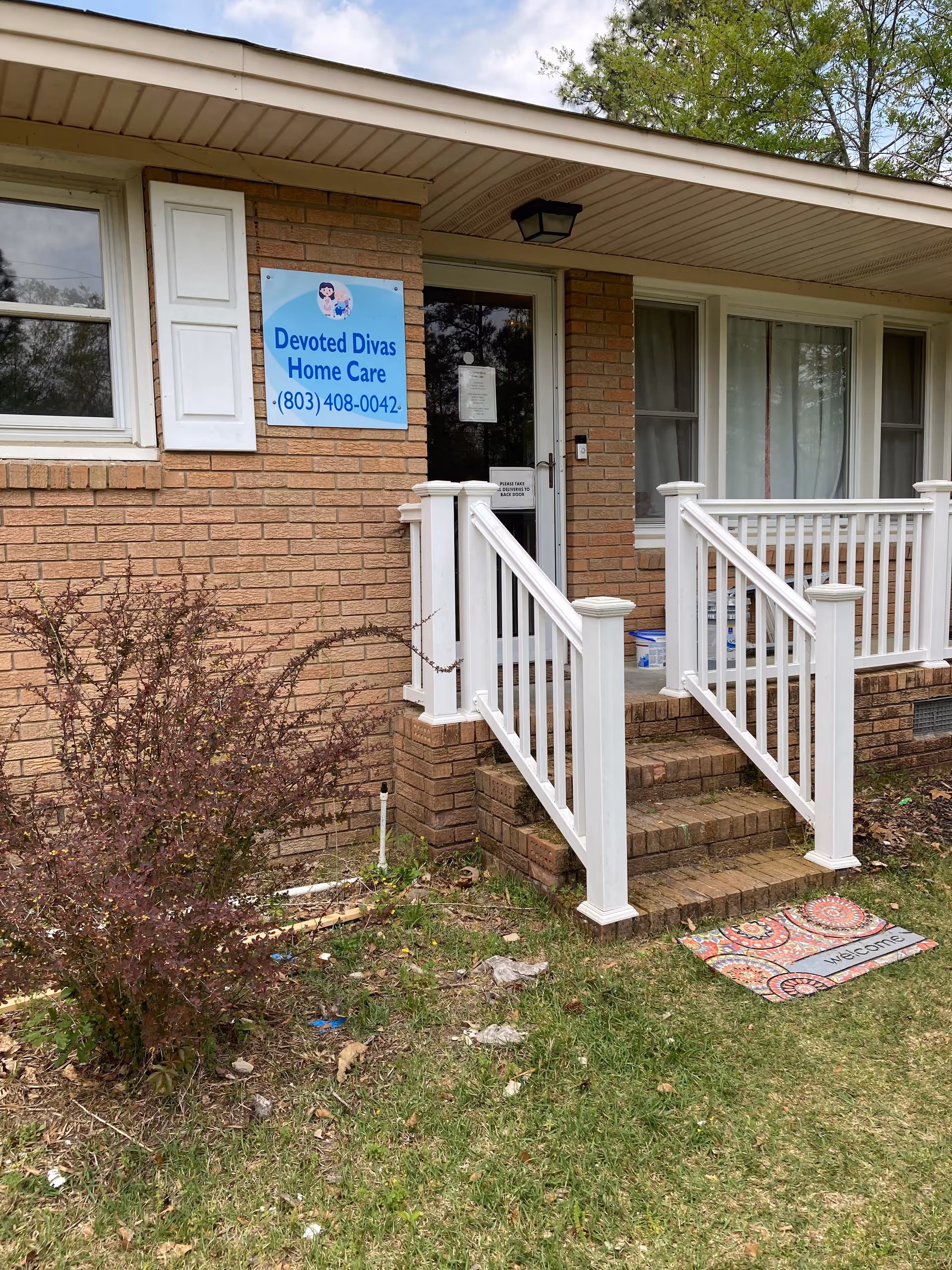 Brick building entrance with white railing, steps, and a blue sign for Devoted Divas Home Care.