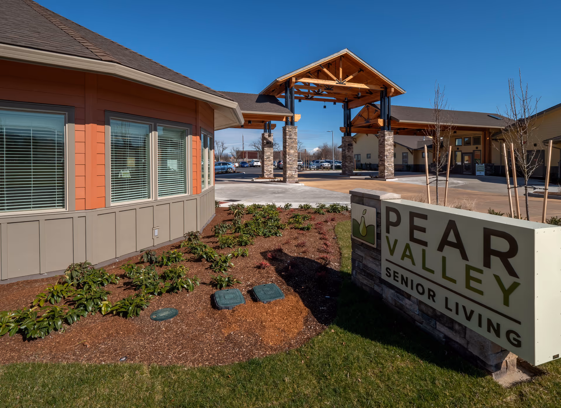 Exterior view of Pear Valley Senior Living facility showing the building entrance with a covered drop-off area supported by stone pillars, landscaped garden beds, and a large sign with the facility name in the foreground under a clear blue sky.