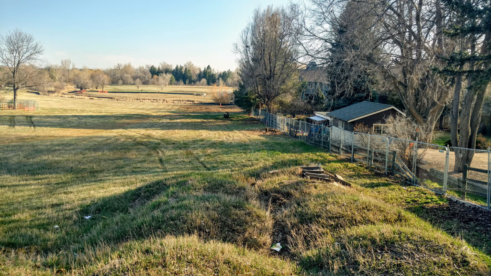 A wide outdoor field with a fence, trees, and a small shed beside a building.