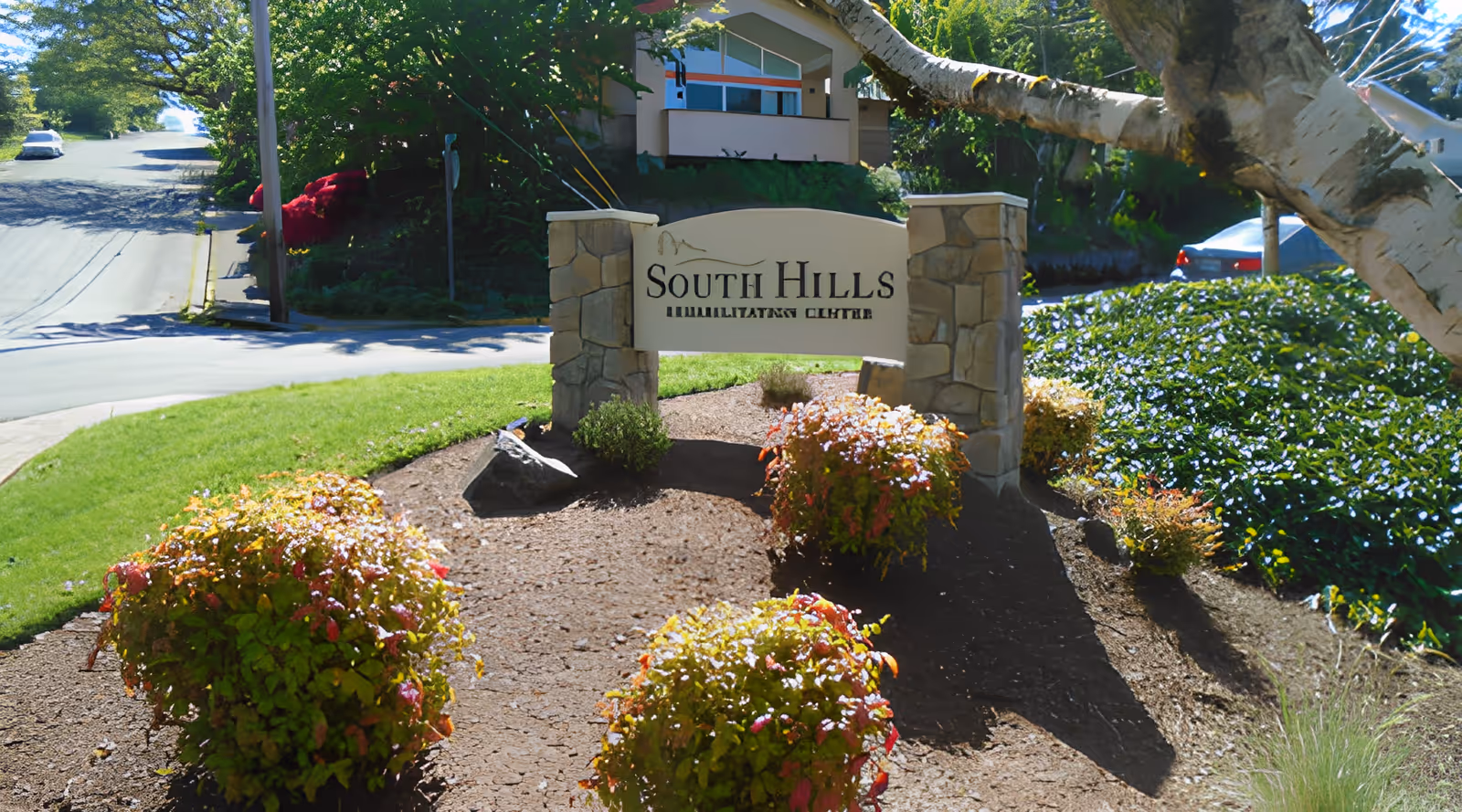 Stone entrance sign reading "South Hills Rehabilitation Center" surrounded by landscaped bushes and trees with a building and street in the background.
