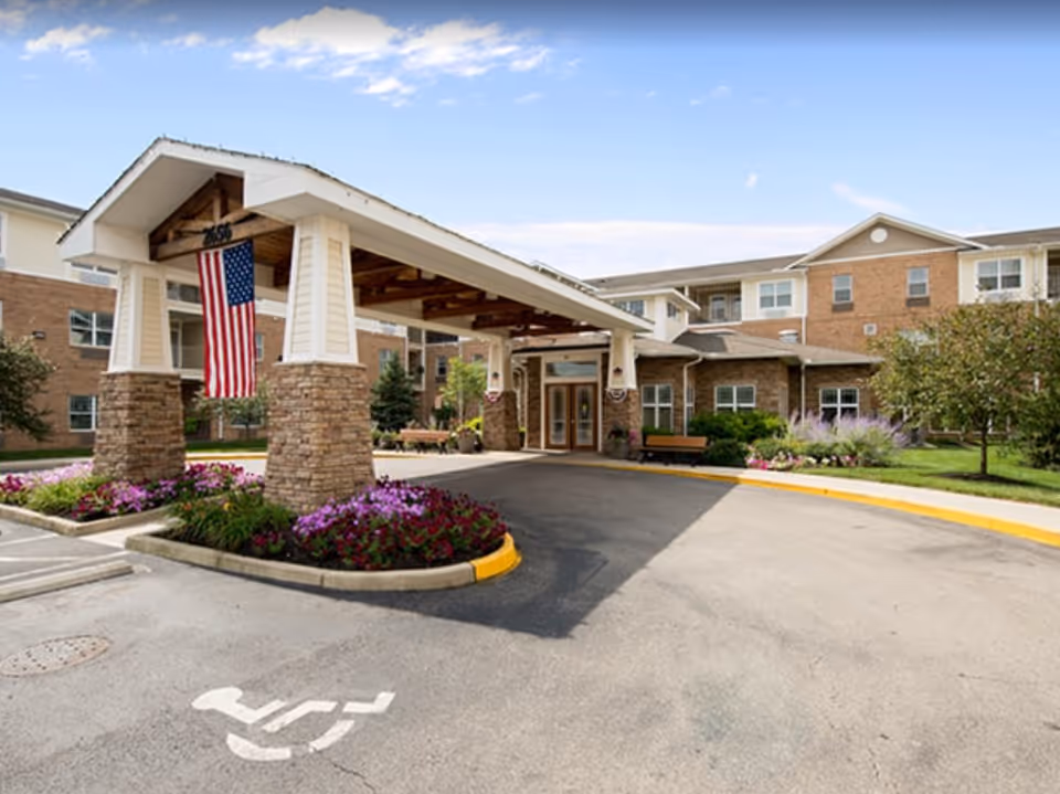 Covered porte-cochere entrance of a brick senior living building with an American flag, benches, and flower beds.