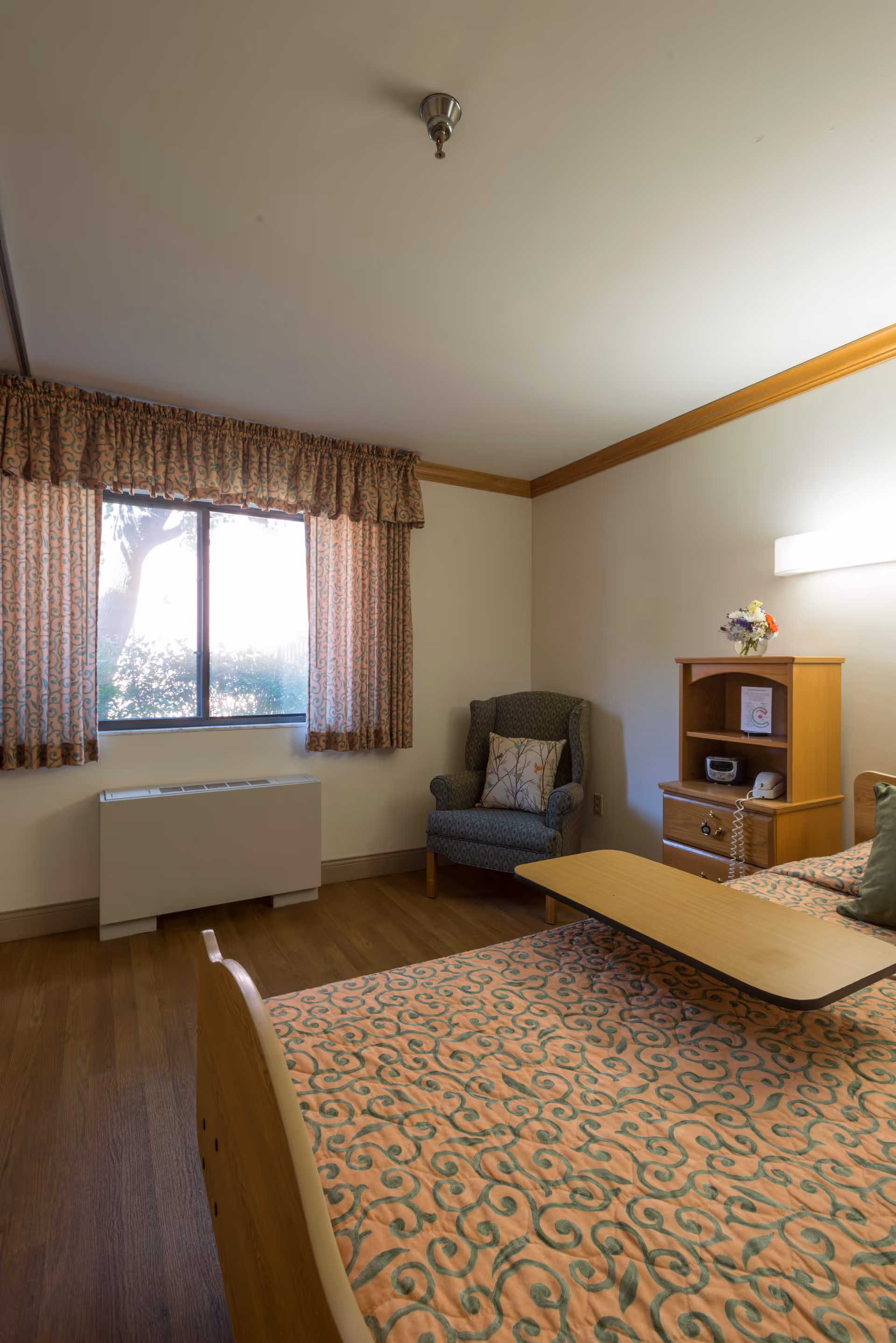 A senior living facility bedroom with a bed covered in a patterned bedspread, a wooden bedside table with a telephone and a small flower arrangement, a cushioned armchair with a decorative pillow, a window with patterned curtains, and a wooden floor.