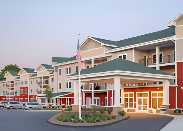 Exterior view of a multi-story senior living facility with beige and red walls, green roofs, and a covered entrance. An American flag is displayed on a flagpole in front of the building, with landscaping and a driveway visible.