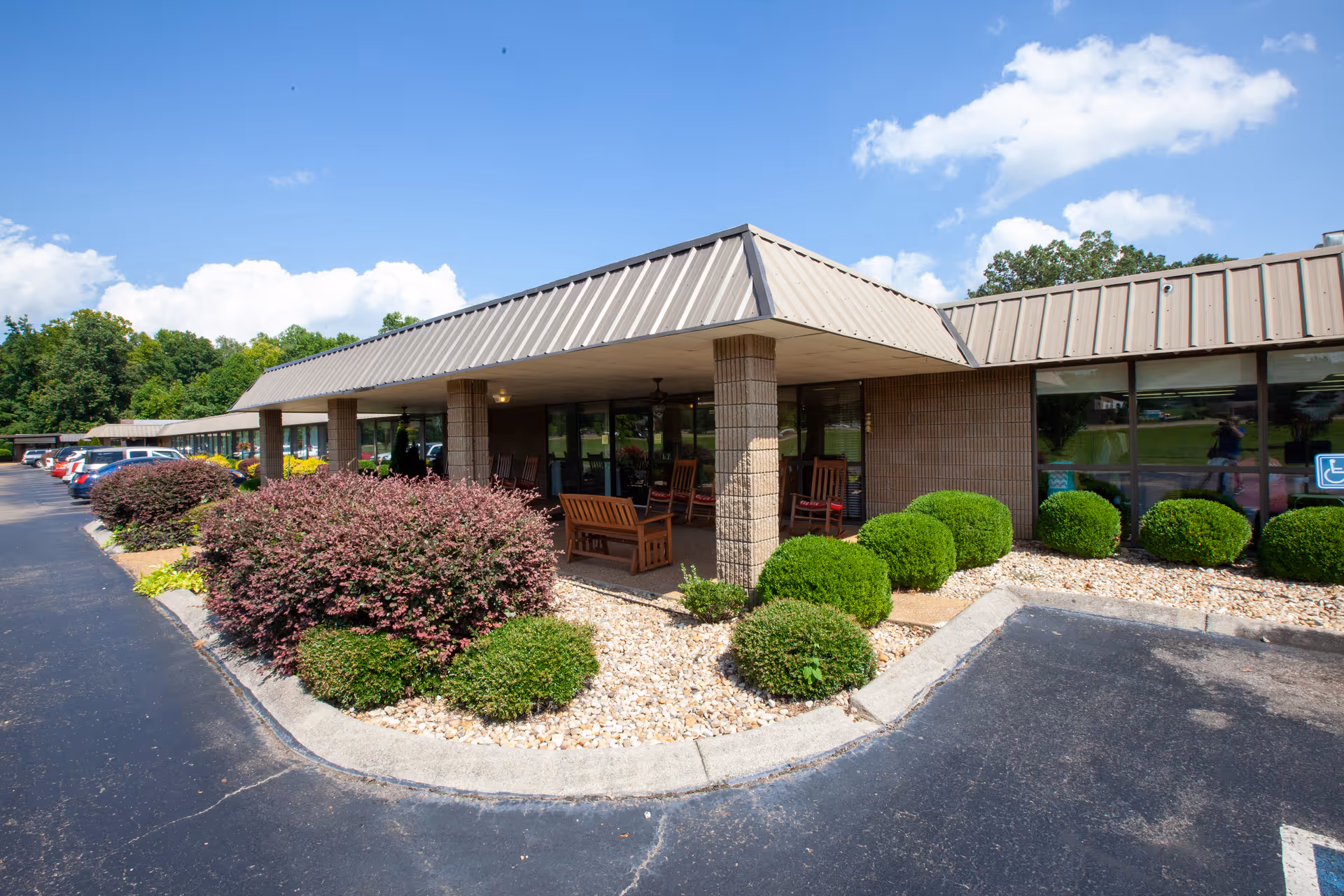 Front entrance of a single-story senior living facility with a covered porch, rocking chairs, and landscaped shrubs.