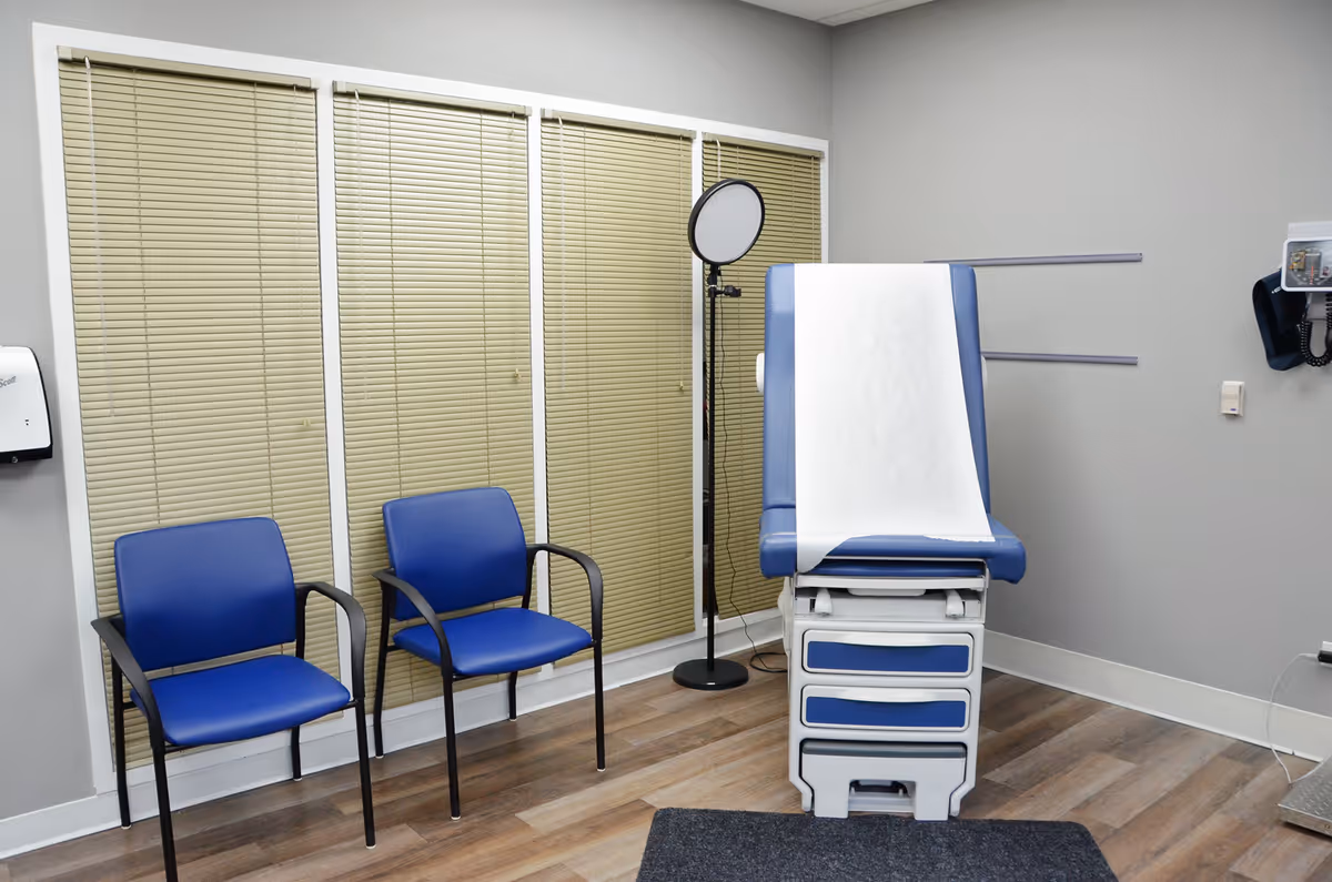 A clinic exam room with two blue chairs, a medical exam table, and blinds.