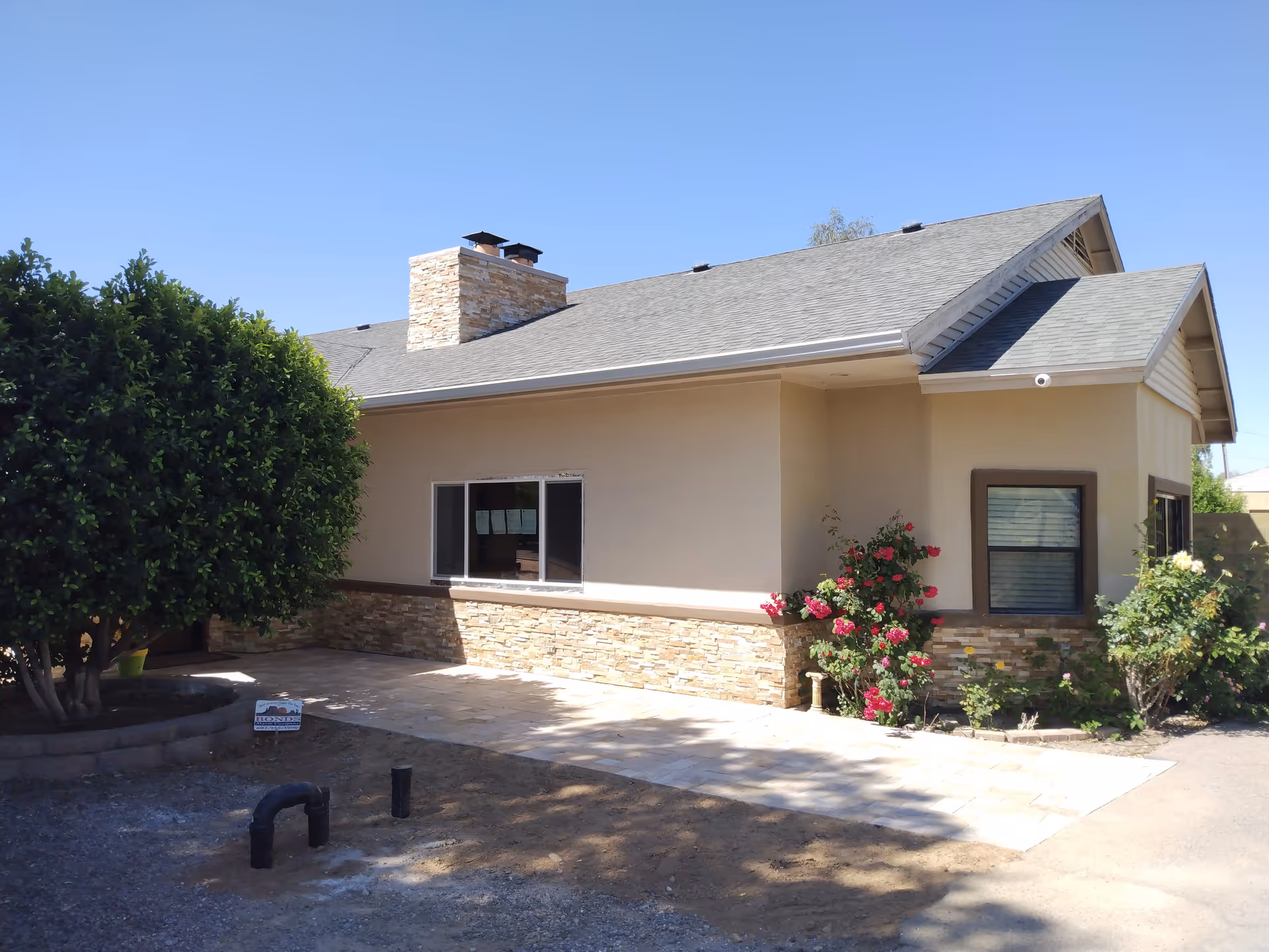 Exterior view of a single-story building with beige walls and a gray shingled roof. The building features a stone chimney and stone accents along the lower part of the walls. There are two windows visible, surrounded by landscaping including a large bush on the left and flowering plants near the right corner. The sky is clear and blue.