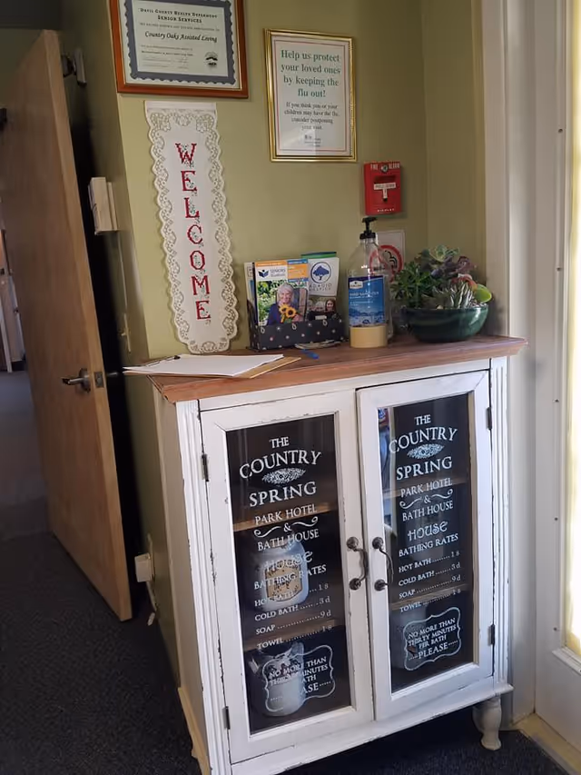 Interior hallway corner with white cabinet, Country Spring signage, plants, notices, and a welcome sign.