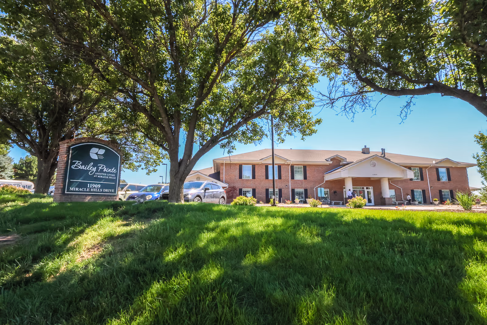 Exterior view of Bailey Pointe at Miracle Hills, a two-story brick assisted living facility with a covered entrance. The building is surrounded by green grass, trees, and a clear blue sky. A sign in front reads 'Bailey Pointe Assisted Living at Miracle Hills 11909 Miracle Hills Drive.'