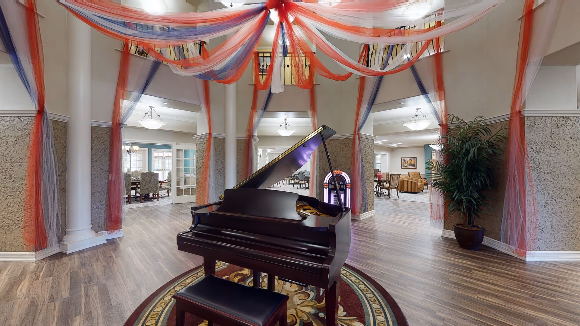 Senior living facility lobby with a grand piano beneath red and blue draped ceiling decorations and nearby seating areas.