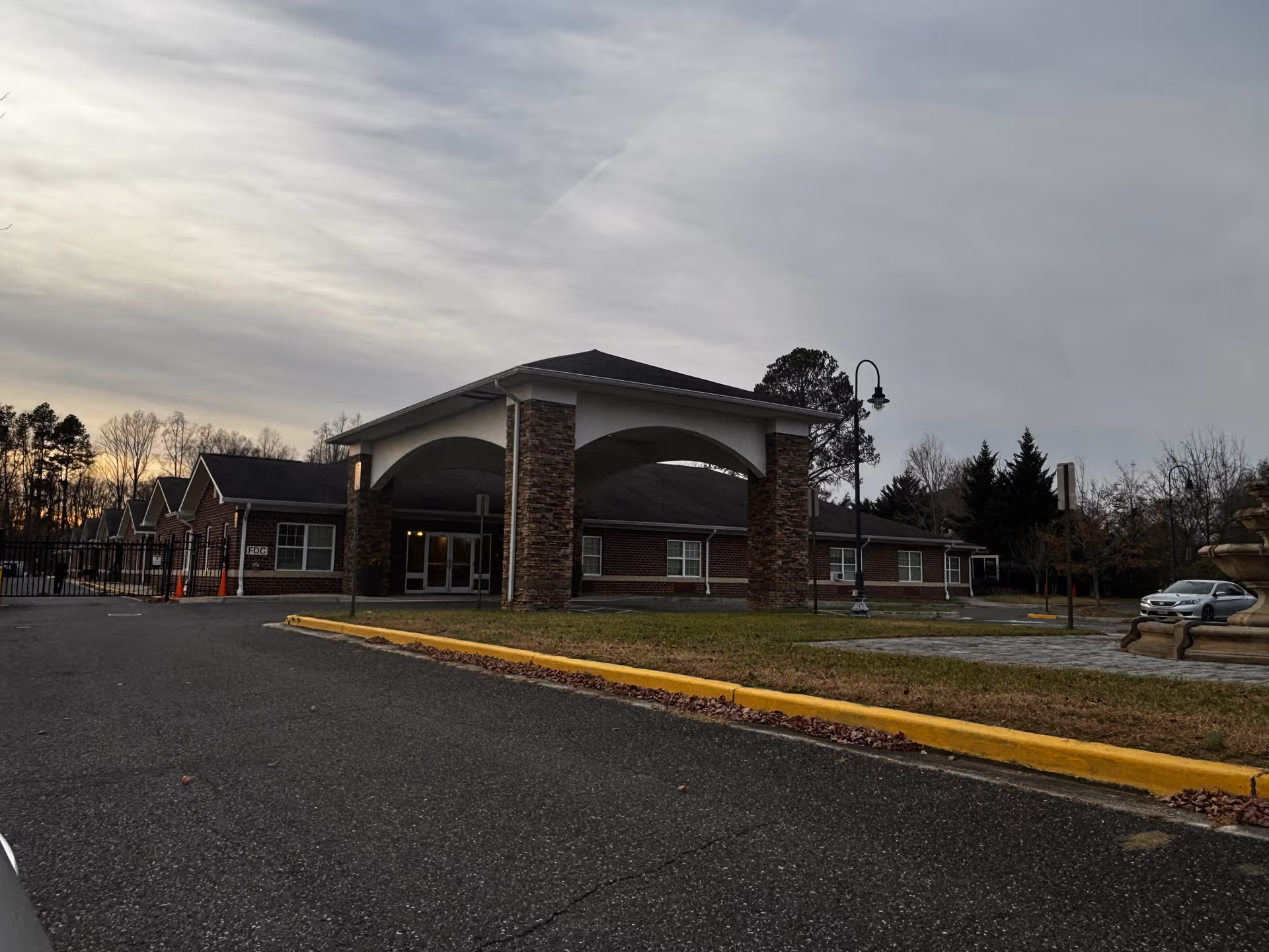 Exterior view of a single-story brick building with a covered entrance supported by stone pillars, a paved driveway, a street lamp, and a car parked on the right side. Trees and a cloudy sky are visible in the background.