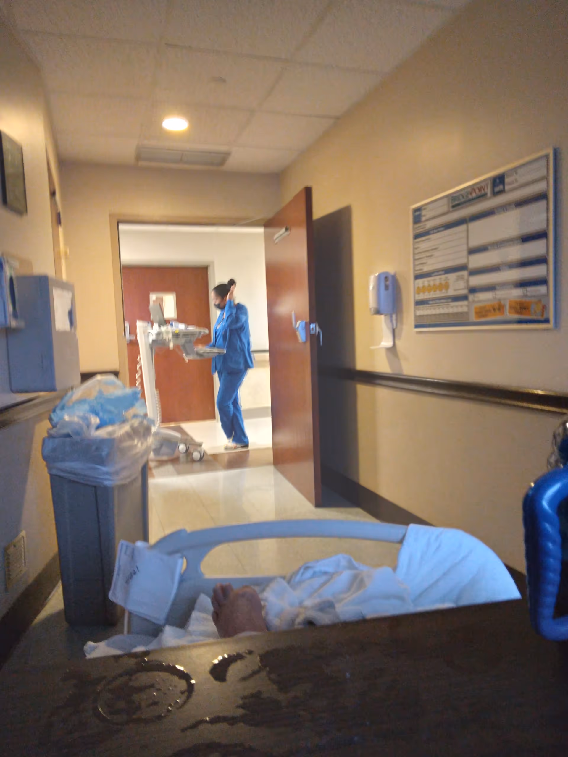 A healthcare worker in blue scrubs pushes a cart down a hospital corridor beside a bed.