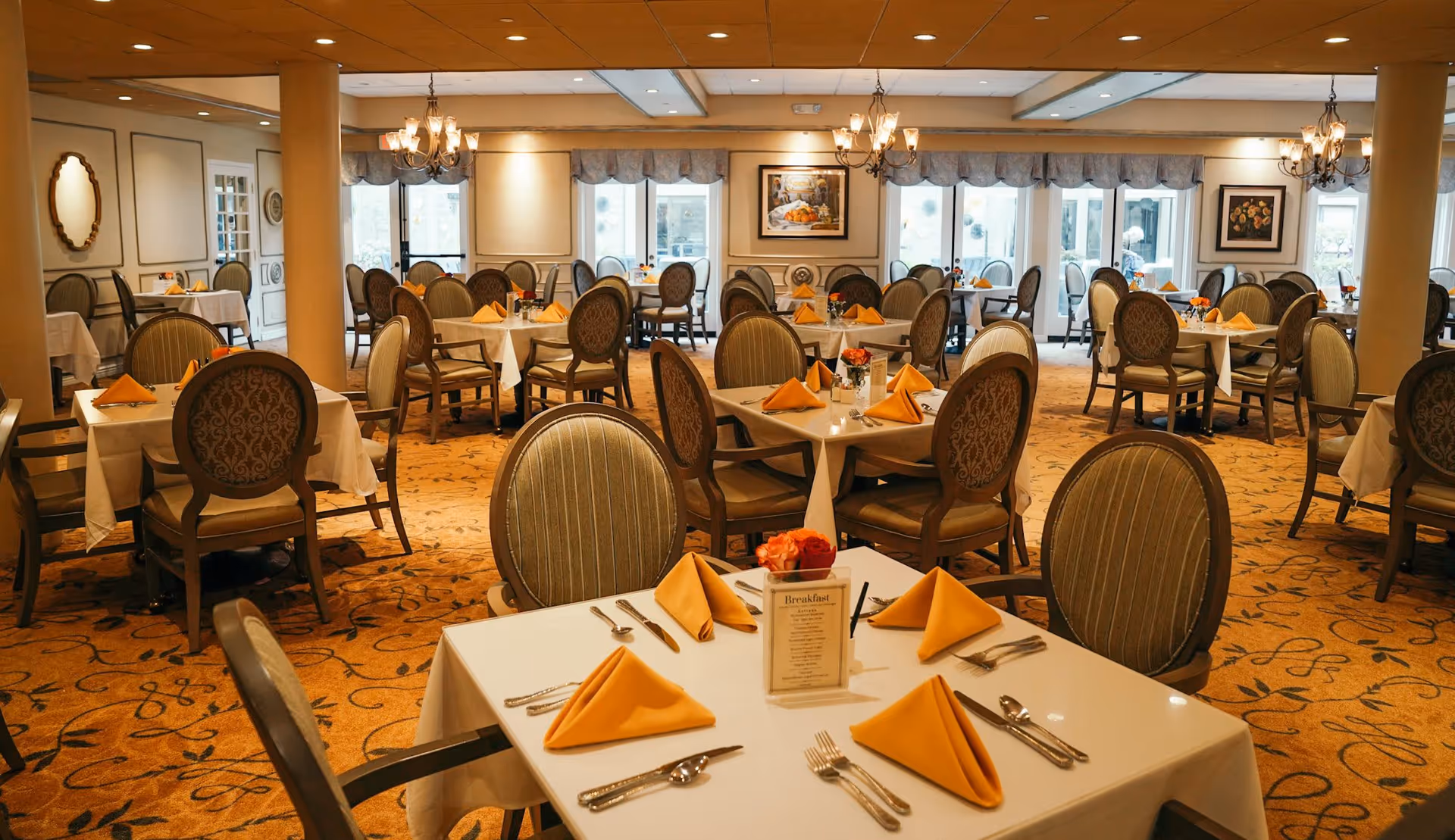Formal dining room with neatly set tables and upholstered chairs, yellow folded napkins, chandeliers, and large windows.