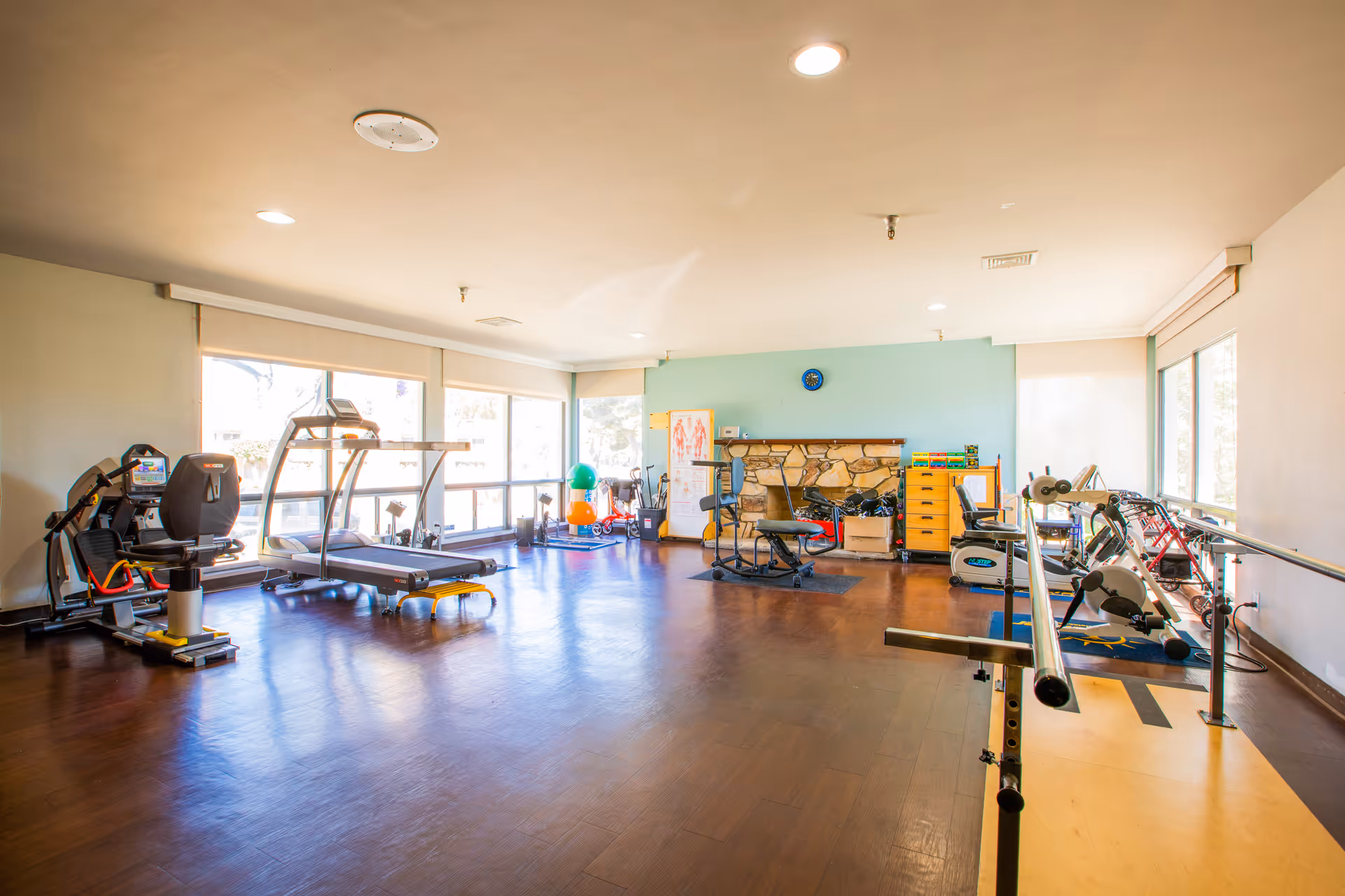 A spacious exercise room with various fitness equipment including a recumbent bike, treadmill, stationary bike, and parallel bars for walking support. The room has large windows allowing natural light, a light blue accent wall with a clock, and a wooden floor.