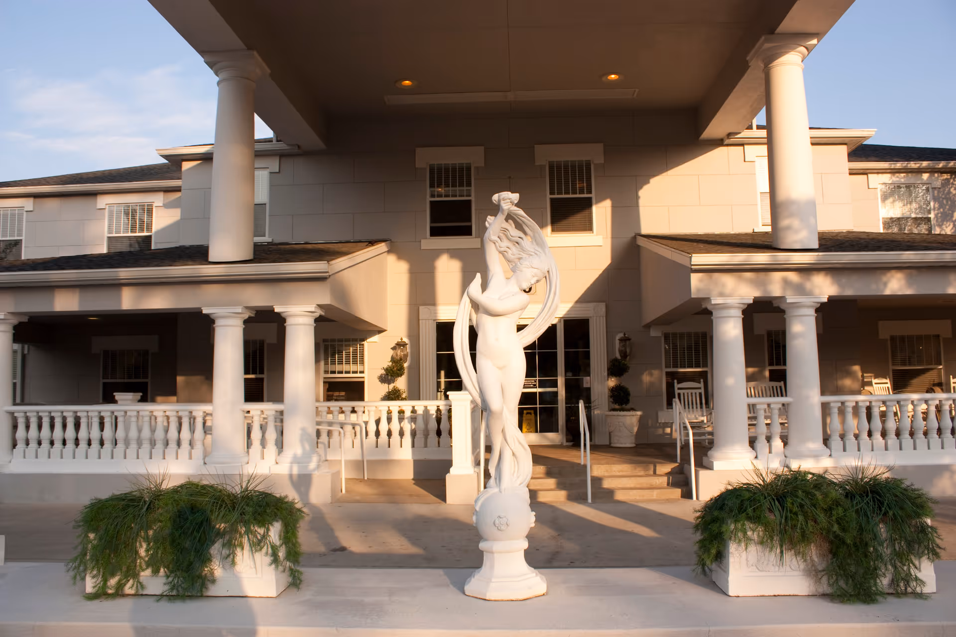 Front entrance of a senior living facility with white columns and railings, featuring a white statue of a woman in the center and two planters with greenery on either side of the walkway.