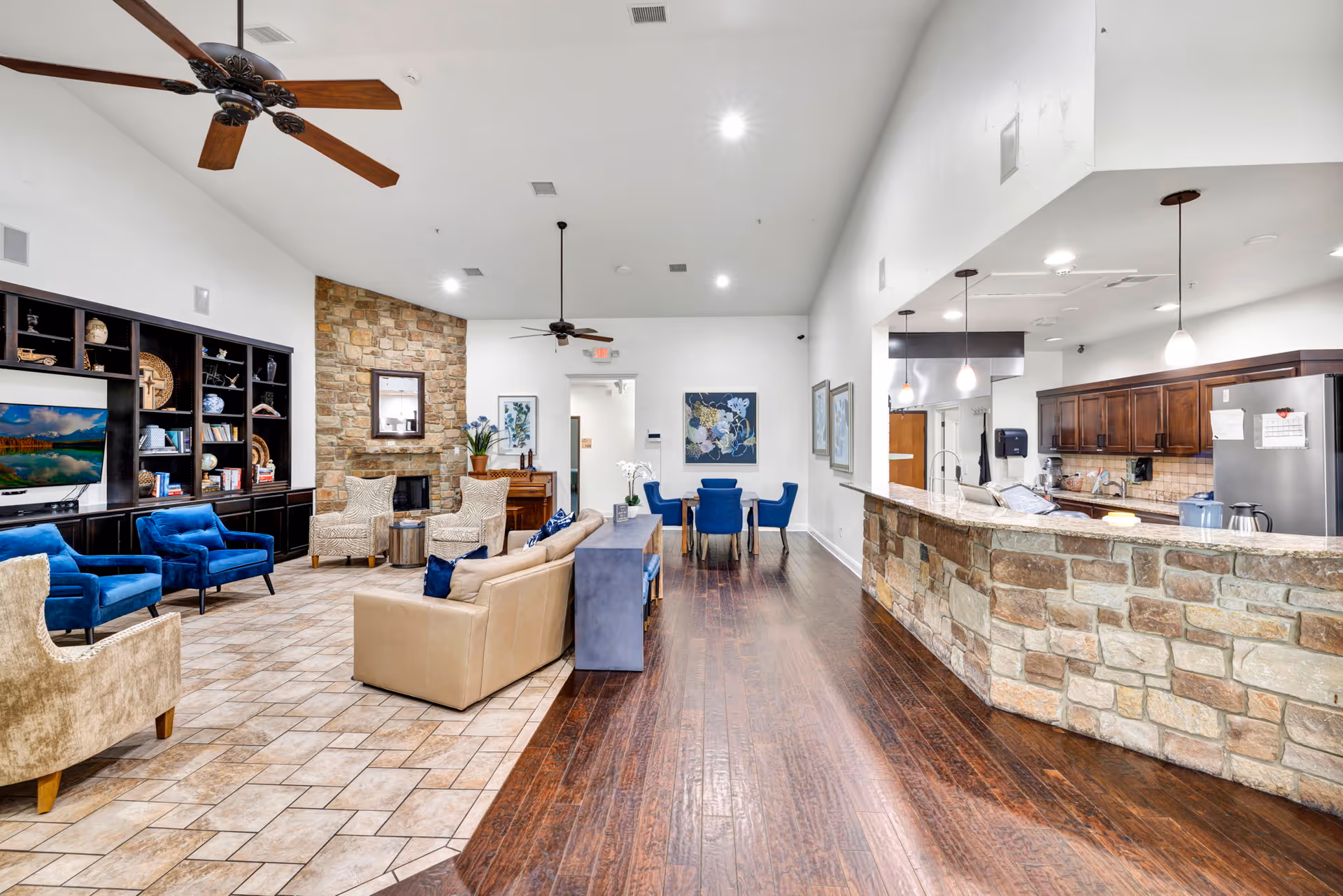 Spacious assisted living common area with lounge seating, a stone fireplace, and an open kitchen/bar.